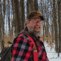 A man wearing a cap and plaid jacket in the woods.